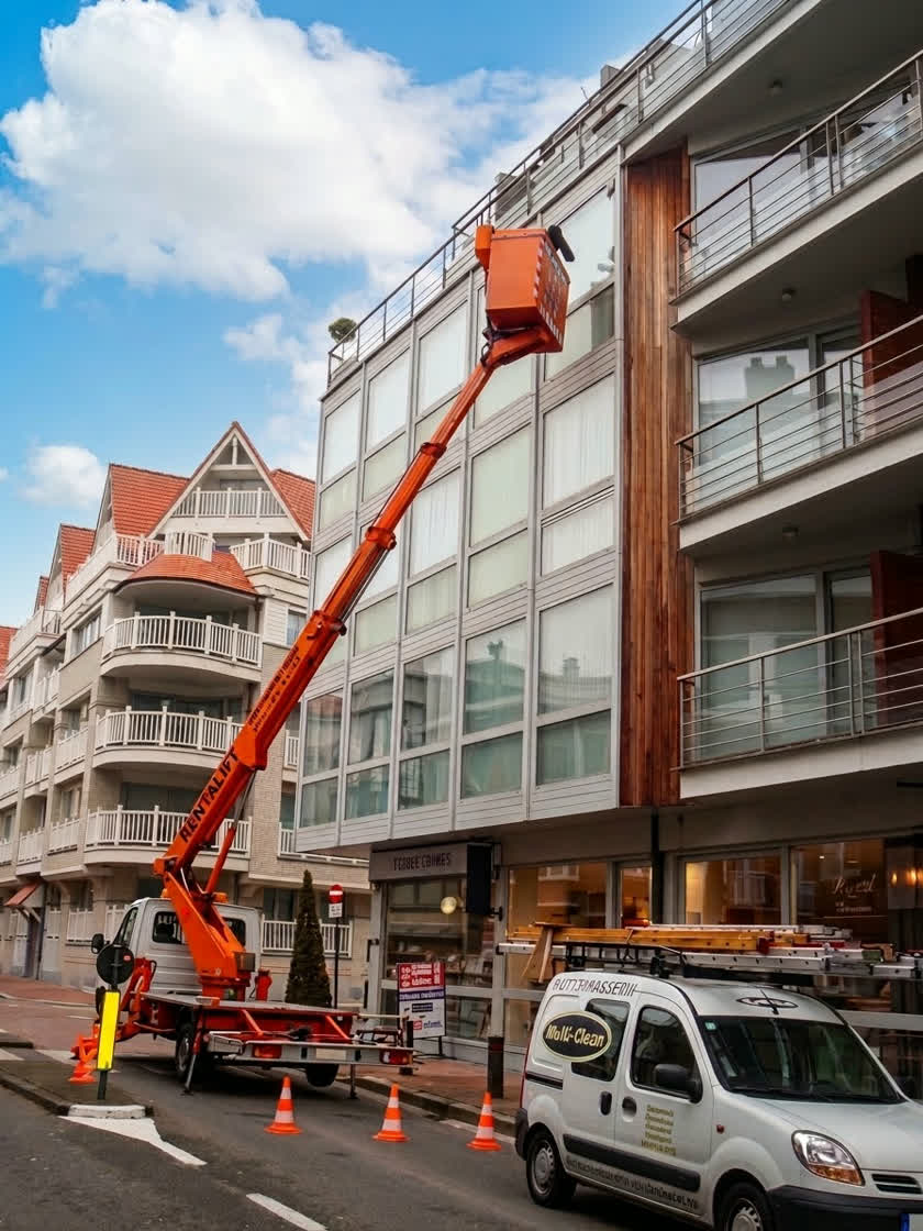 Een hoogtewerker met uitgeschoven arm reikt tot de bovenste verdieping van een appartementsgebouw in een straat aan de kust. Onderaan staat de bestelwagen van Multi-Clean met verkeerskegels errond.