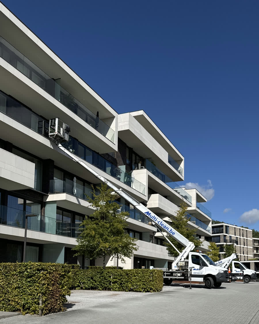 De hoogtewerker van Multi-Clean staat op een eigen bestelwagen voor een modern appartementsgebouw. De arm is uitgeschoven tot de bovenste verdieping en draagt het webadres multi-clean.be. De mand hangt aan een raam van het bovenste appartement.